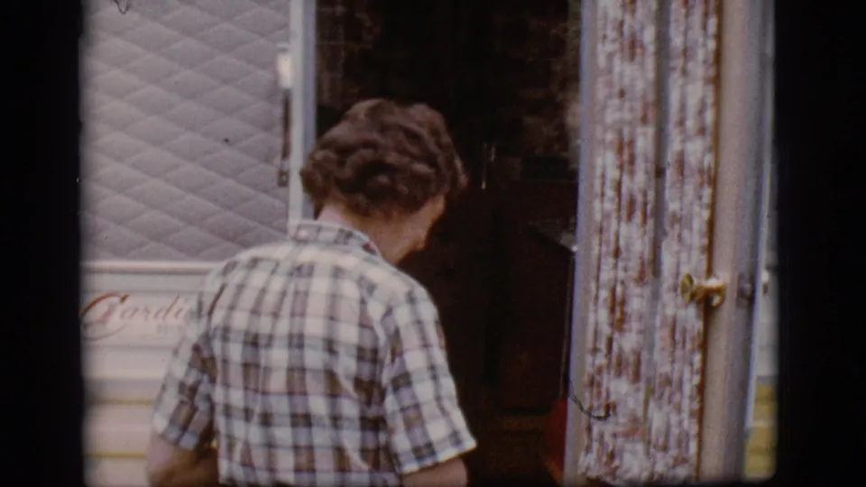Seen from behind, the plaid-shirted woman leans toward the camper interior, framed by weathered trim and pale siding.
