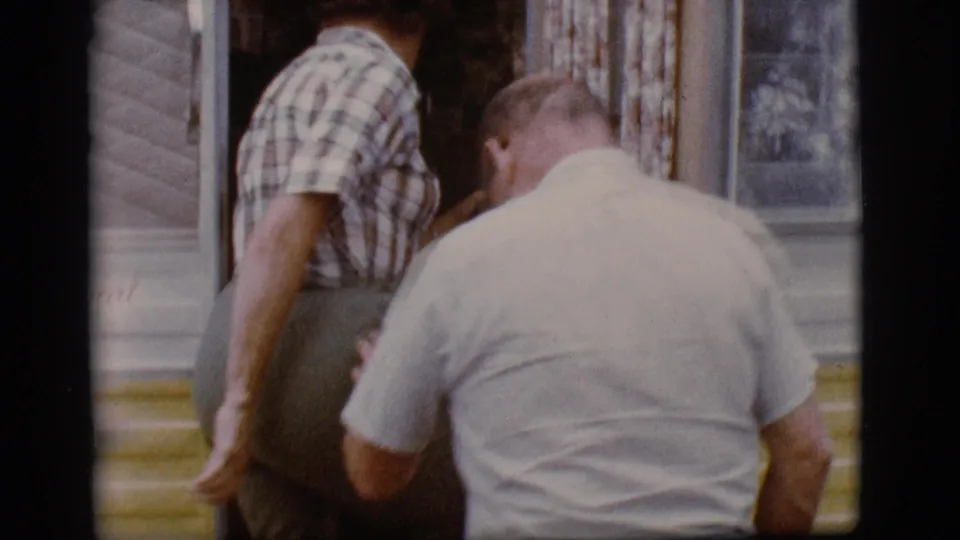 A man in a light shirt follows the woman in plaid toward the open camper door in 1968 Flagstaff vacation footage.