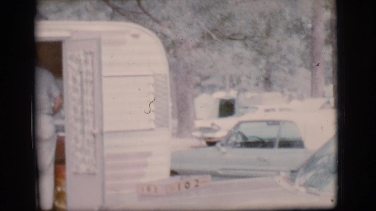 A 1960s travel trailer doorway is seen at a campground, with parked cars and another camper visible beyond it.