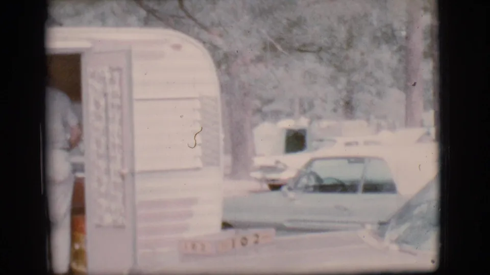 A 1960s travel trailer doorway is seen at a campground, with parked cars and another camper visible beyond it.