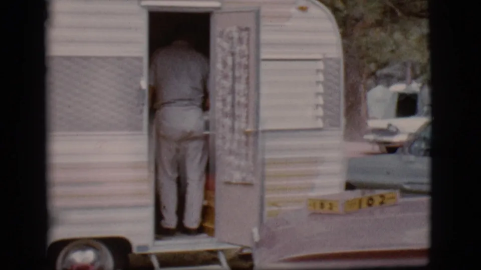The man is mostly inside the trailer doorway while a parked car and another camper sit in the campground background.