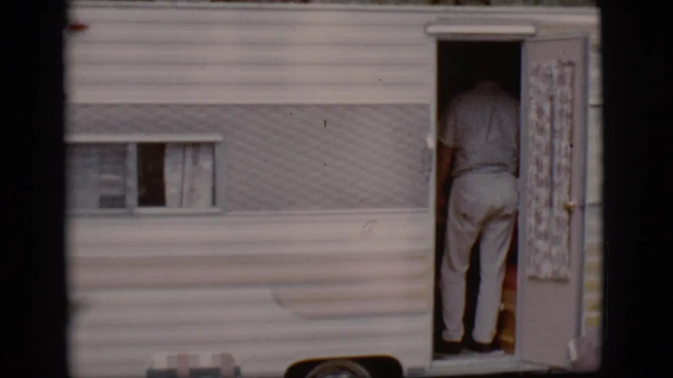 A man in light trousers stands inside the open doorway of a 1960s travel trailer, seen from behind in Flagstaff footage.