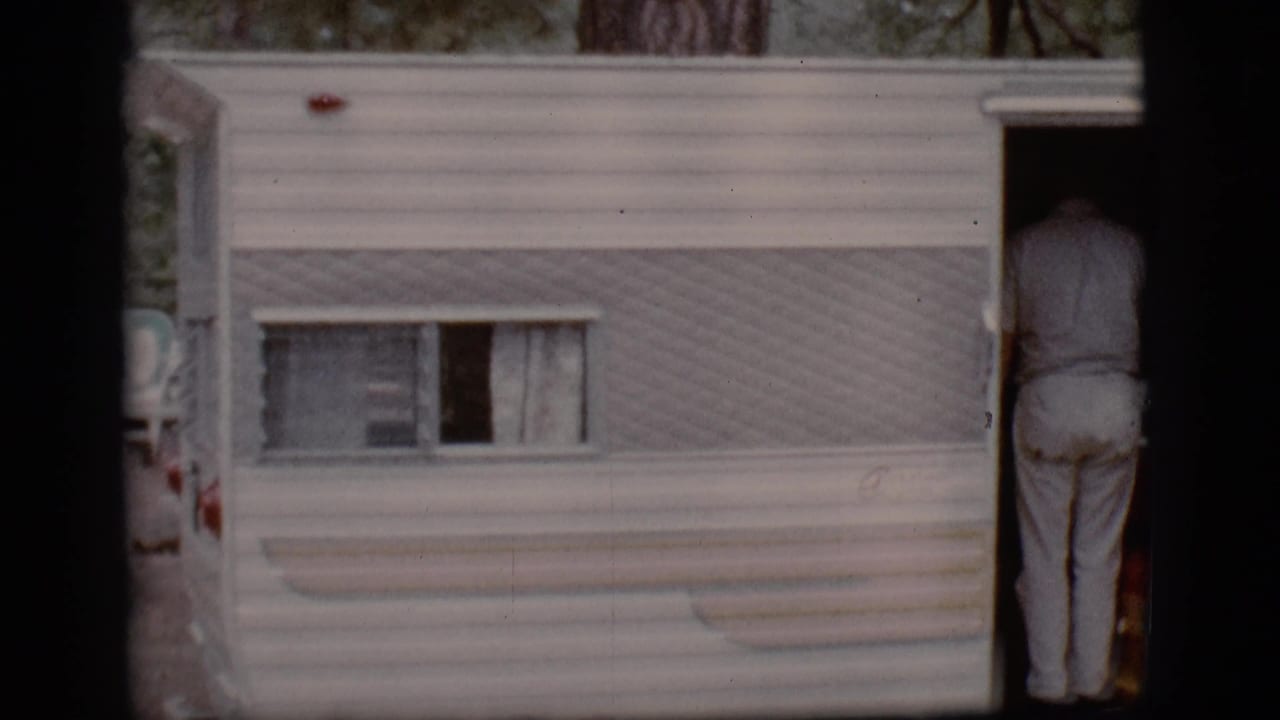 White camper trailer with a person standing in the open doorway at a wooded campground.
