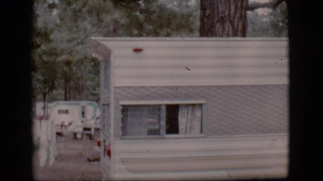 Side of a white camper trailer with a curtained window and campground trailers in the background.