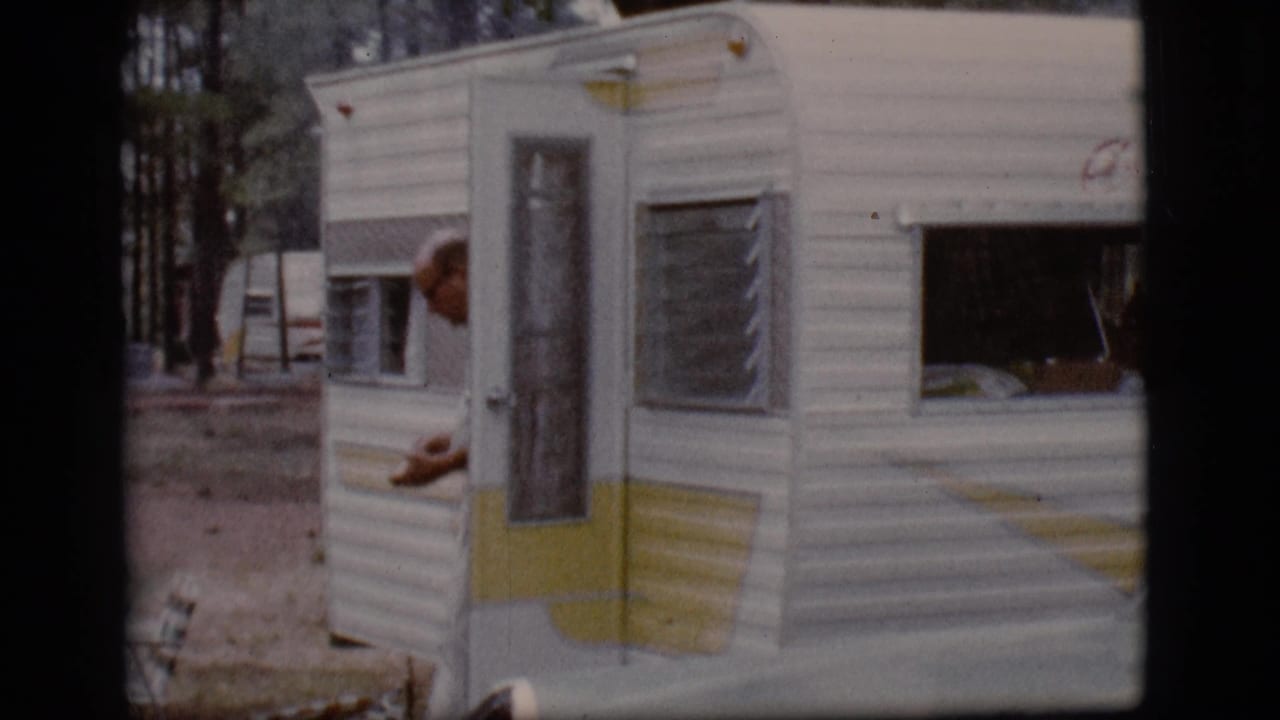 Man partly visible beside a white and yellow trailer in a pine campground.