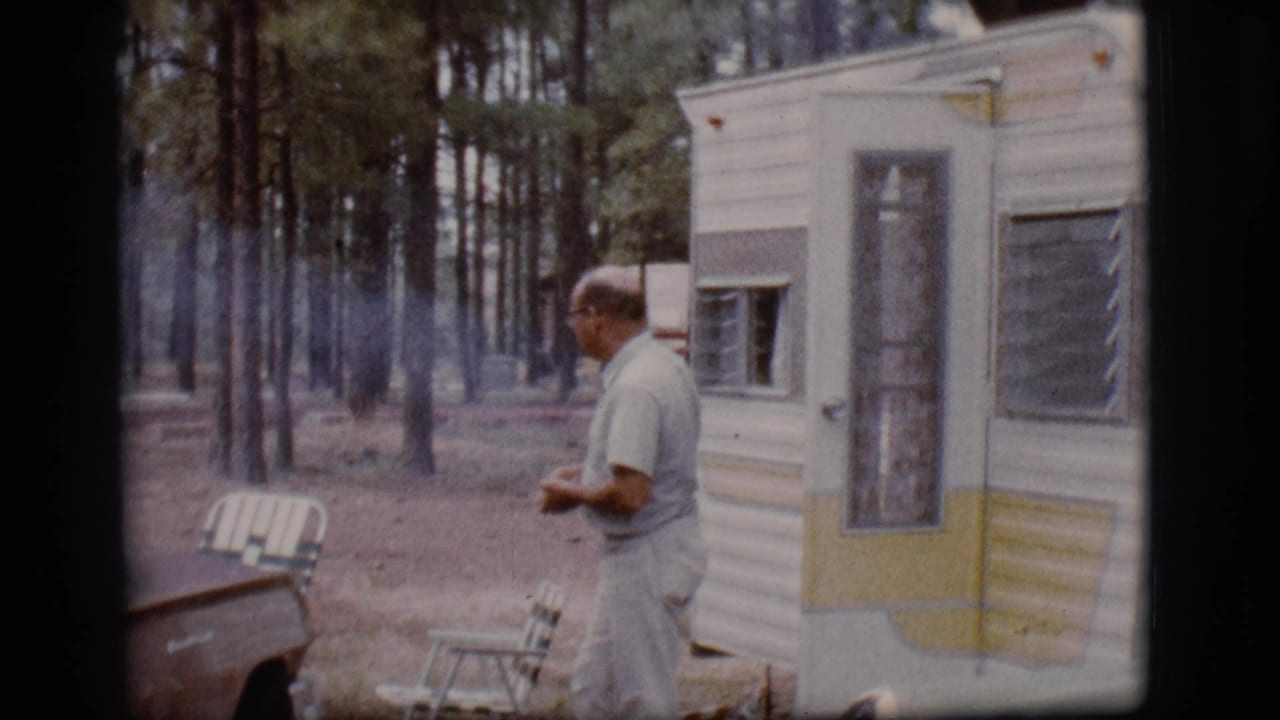 Man in light clothing stands near a trailer with trees and a parked car nearby.