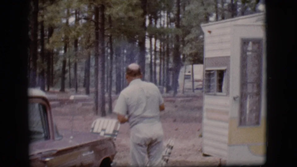 The man walks away from the caravan trailer toward a parked car, with chairs and pine trees visible in the 1968 Flagstaff scene.