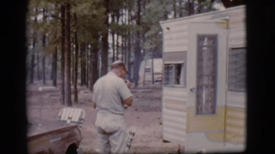 A rear view of the man near the white and yellow trailer, with a parked car, chairs, and tall pines in the Flagstaff campground.