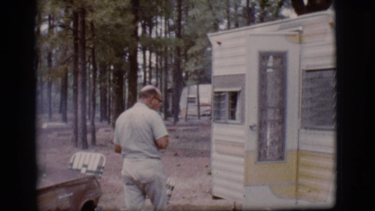 Man walking toward a white and yellow camper trailer in a wooded campsite.