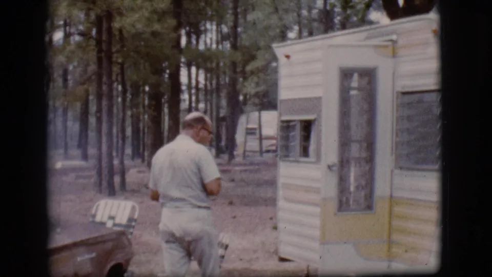 In 1968 Flagstaff, Arizona footage, a man in light clothing approaches a mid-century camper trailer among tall trees.