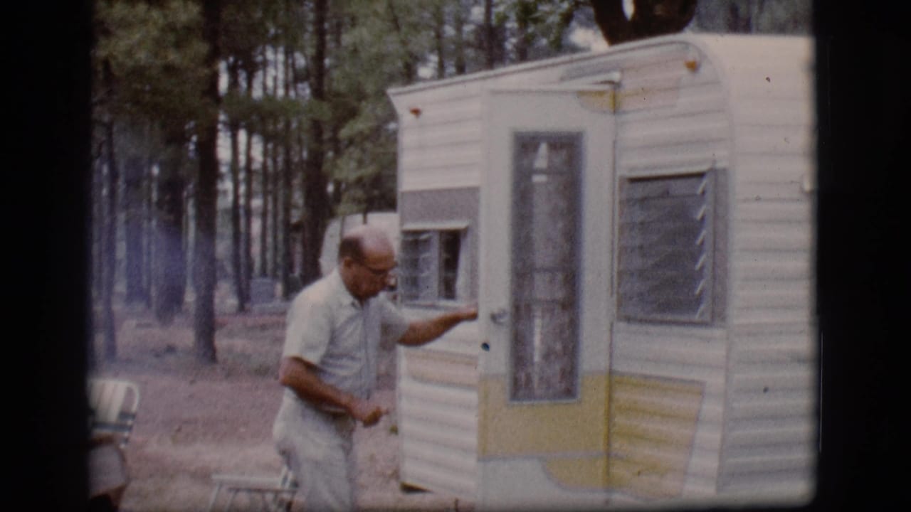 Man standing by the camper trailer door and gesturing toward the camera.
