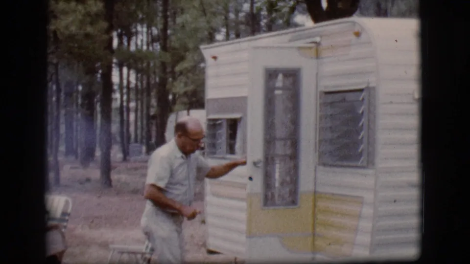 The man stands beside the camper trailer door at the wooded Flagstaff campsite and appears to wave or gesture toward the camera.