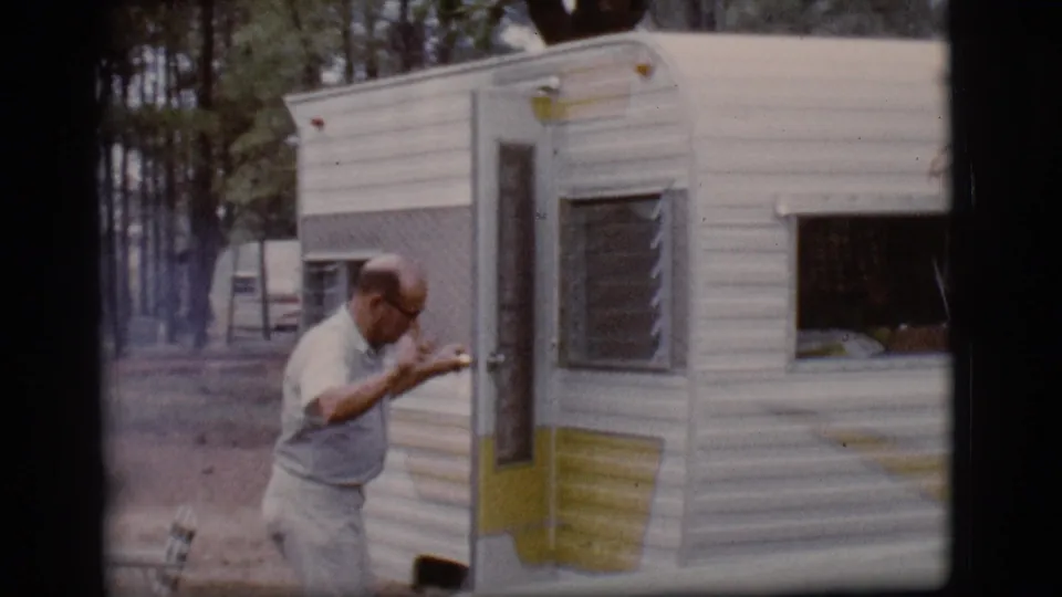 The man moves along the side of the yellow-and-white camper trailer, with louvered windows and pine trees visible behind him.