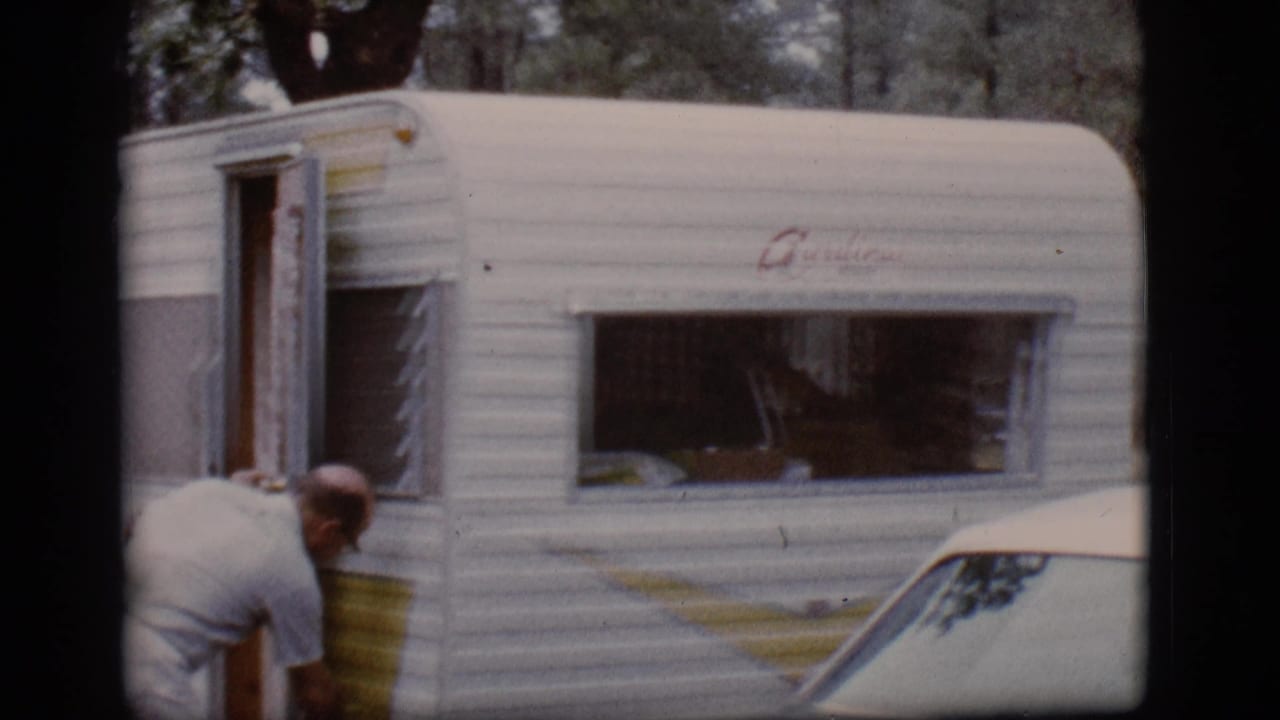 Man bends beside the open door of a white camper trailer in a wooded campsite.