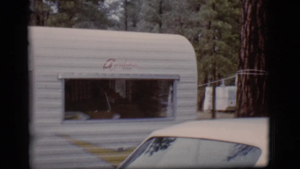 White 1960s camper trailer sits at a wooded Flagstaff campsite, with a partial light-colored car in the foreground.
