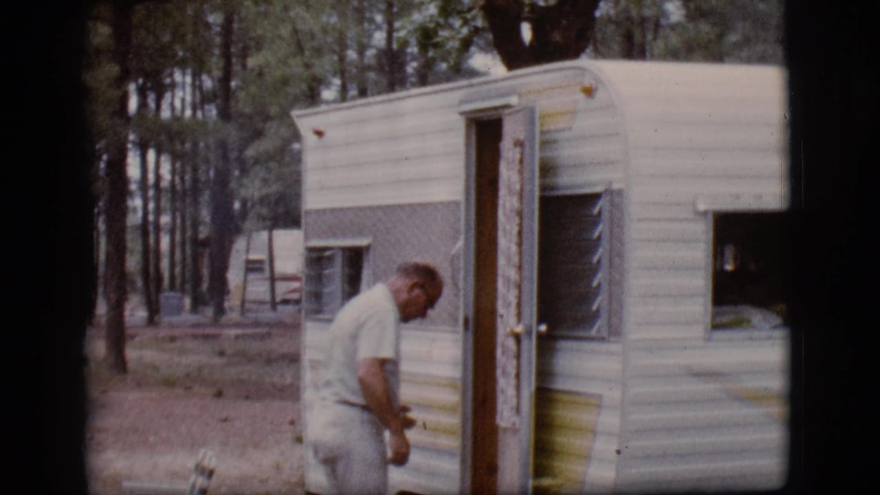Man approaches a small vintage camper trailer at a wooded campsite.