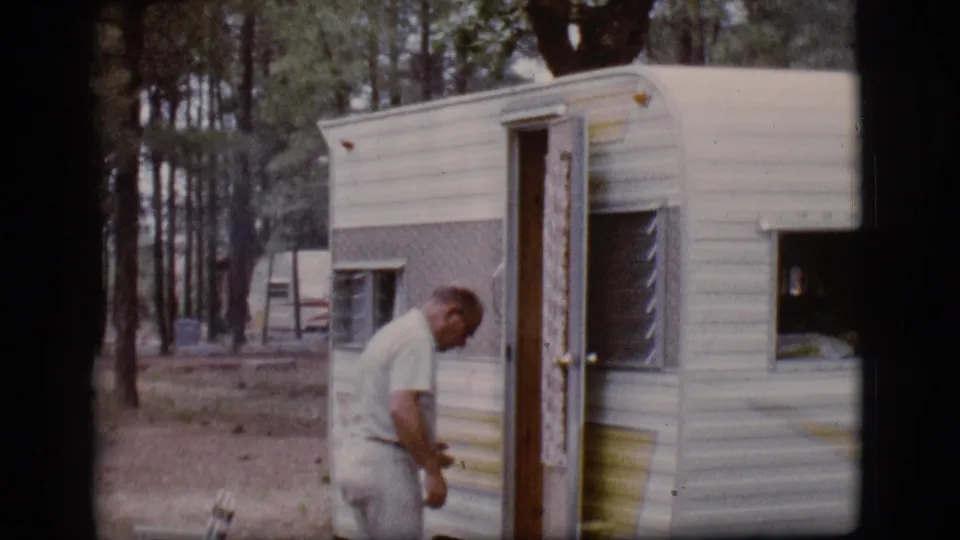 In 1968 Flagstaff, Arizona footage, an adult man stands beside a white and gray camper trailer among pine trees.