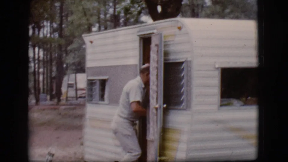 The man enters the open door of the small camper trailer in wooded 1968 Flagstaff campsite footage.