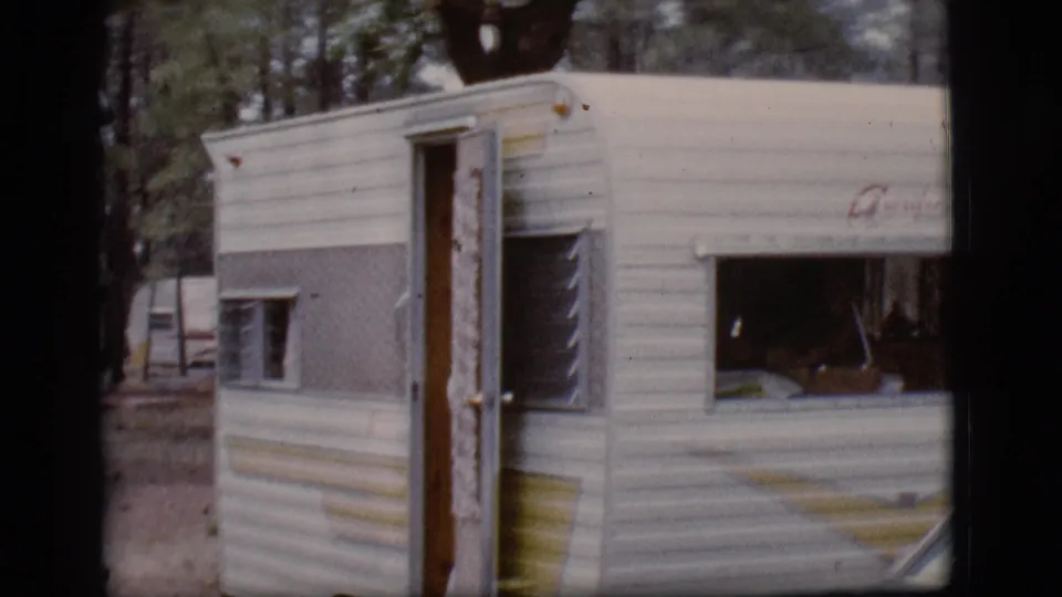 A white and gray 1960s camper trailer sits among trees with its door open and interior partly visible.