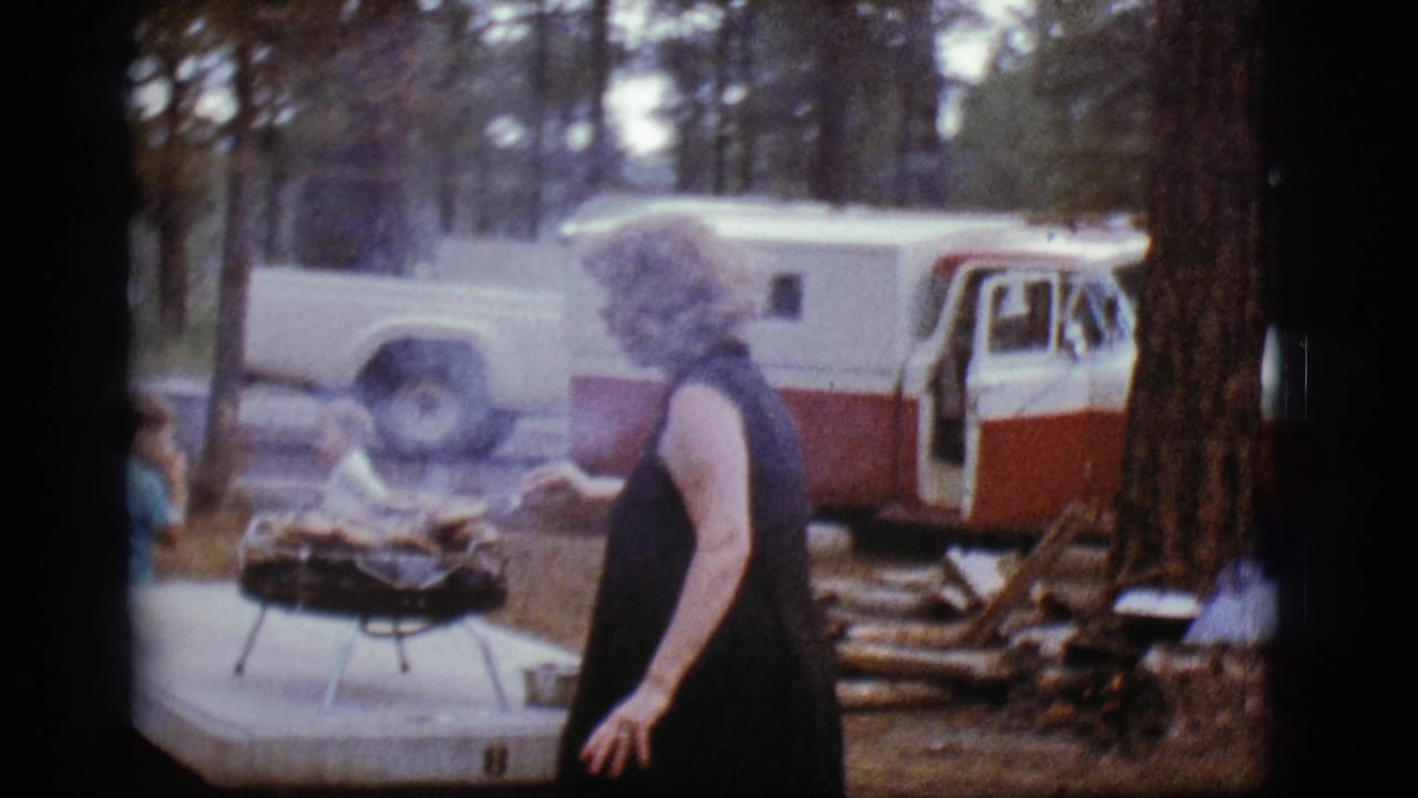Woman beside a smoking portable grill at a wooded campsite with a camper vehicle behind her.