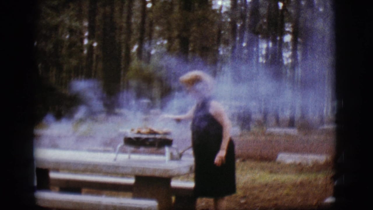 Woman stands near a smoky grill on a picnic table in a pine forest campsite.