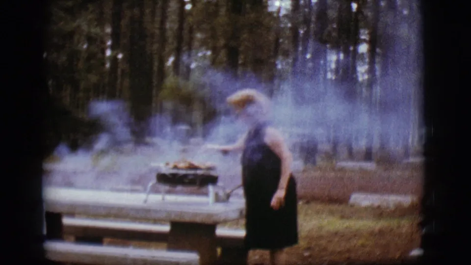 In the 1968 Flagstaff campsite footage, the woman stands beside a smoky portable grill set on a picnic table among tall pines.
