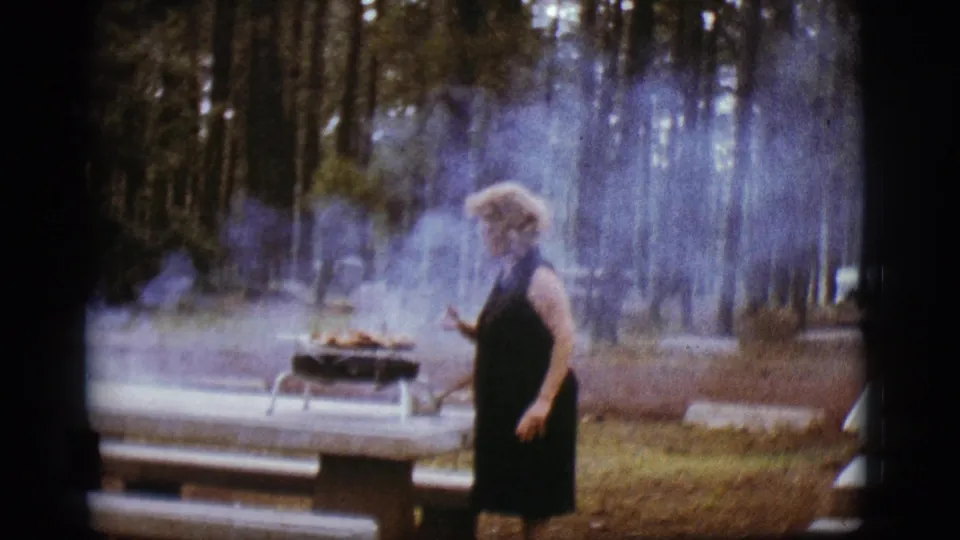 The woman appears to check the food on a smoky portable grill at a Flagstaff pine campsite in 1968 home-movie footage.