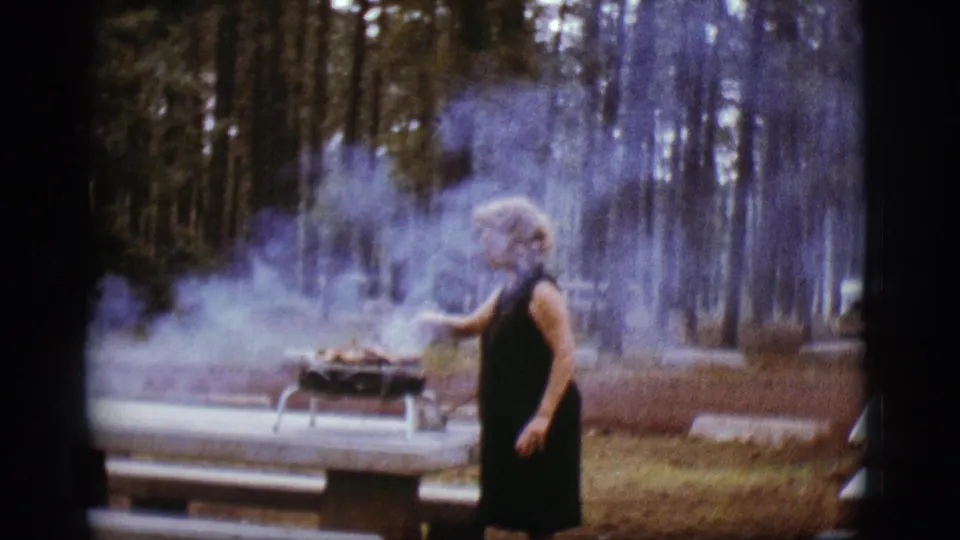 At the wooded Flagstaff campsite, the woman reaches toward the portable barbecue as smoke rises in the 1968 8mm footage.