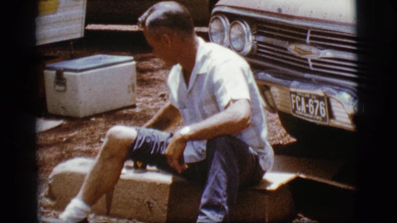 Man looks aside while seated near a classic car and white cooler.