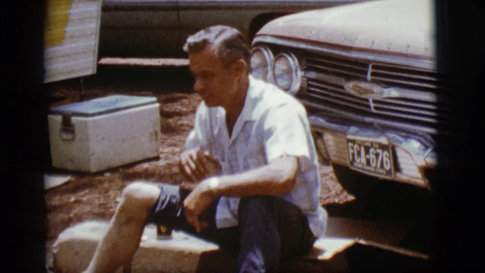 The man sits casually with a small dark drink visible near his knee, beside the Chevrolet front end and cooler.