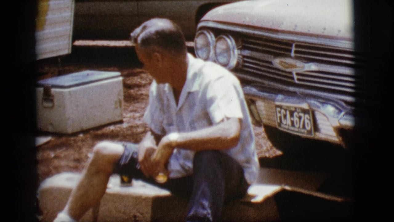 Man seated on a curb beside a Chevrolet, holding a drink near a white cooler.