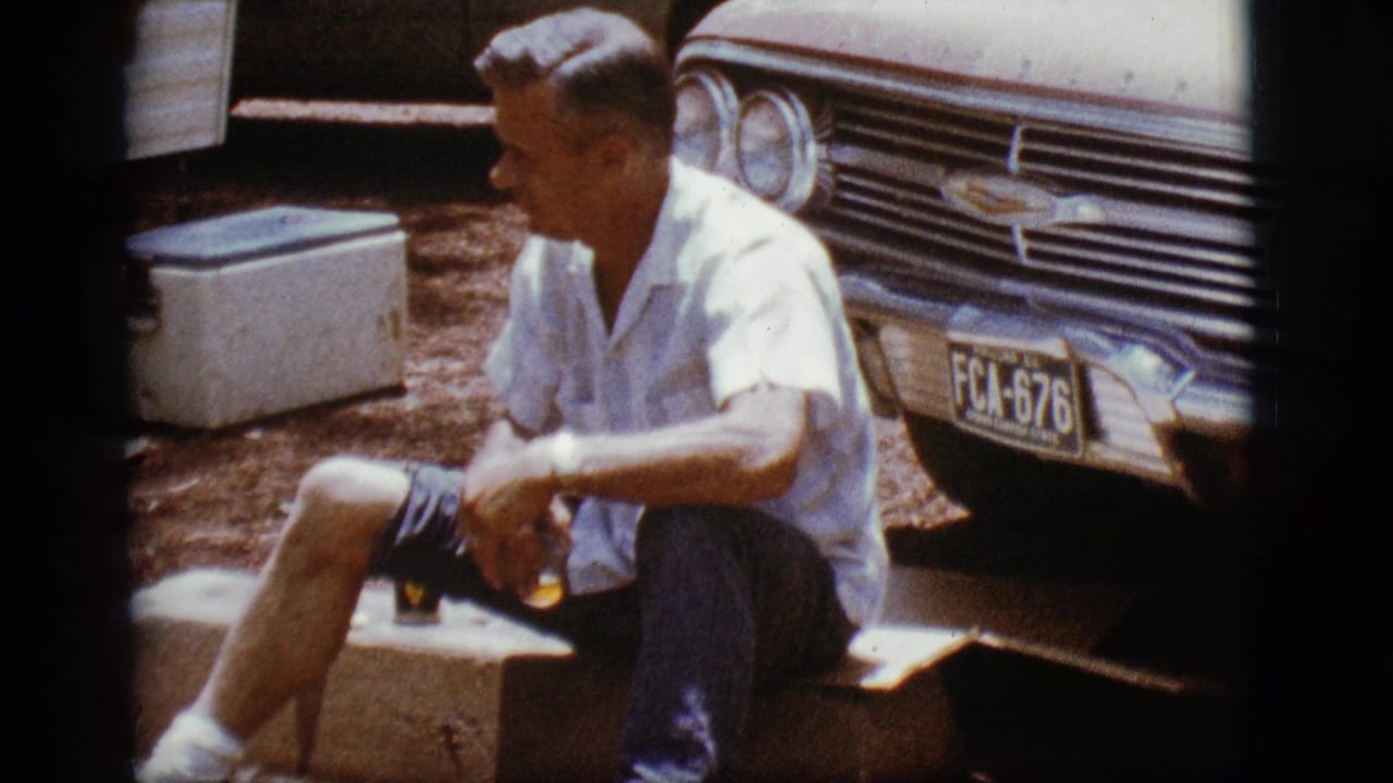 Man sitting beside the front of a Chevrolet, drink in hand, with cooler behind him.