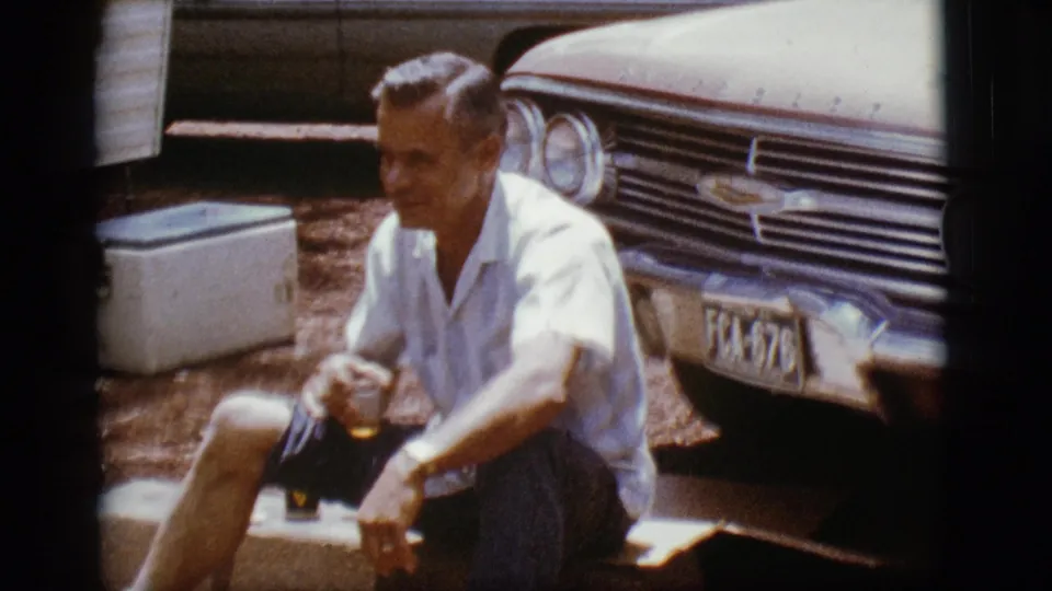 The seated man faces the home-movie camera beside the Chevrolet, with a drink visible near his knee and a cooler behind him.