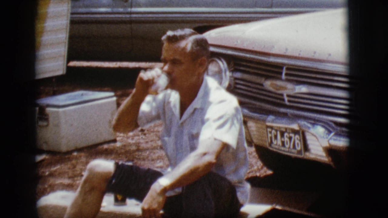 Adult man seated outdoors drinking beside a vintage Chevrolet and white cooler.