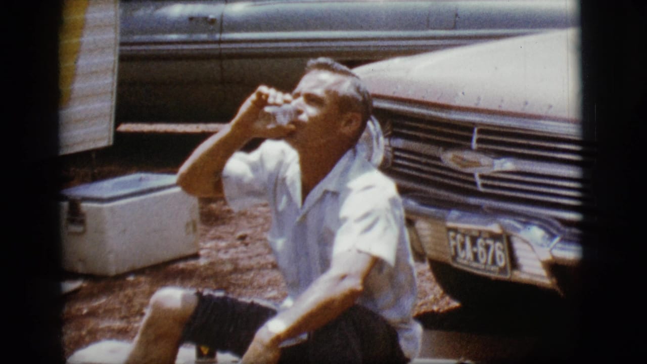 Man raises a glass while seated near a car grille, cooler, and trailer.