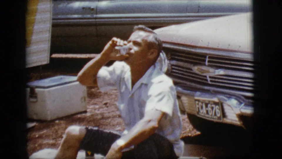The man tilts a glass to drink while seated in a sunlit trailer-park setting in the 1968 Flagstaff home movie.
