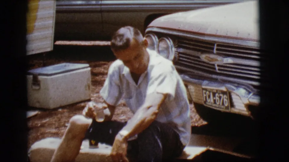 The man lowers his gaze while holding a glass, with the cooler, trailer edge, and Chevrolet front end still visible nearby.
