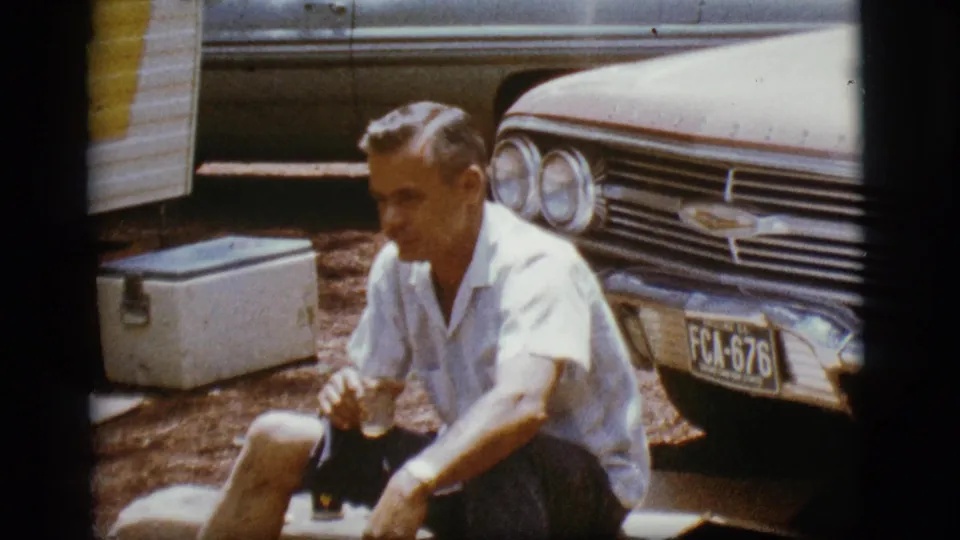 A man remains seated beside the cooler and Chevrolet in the Flagstaff trailer-park scene, with warm 8mm color and visible frame edges.