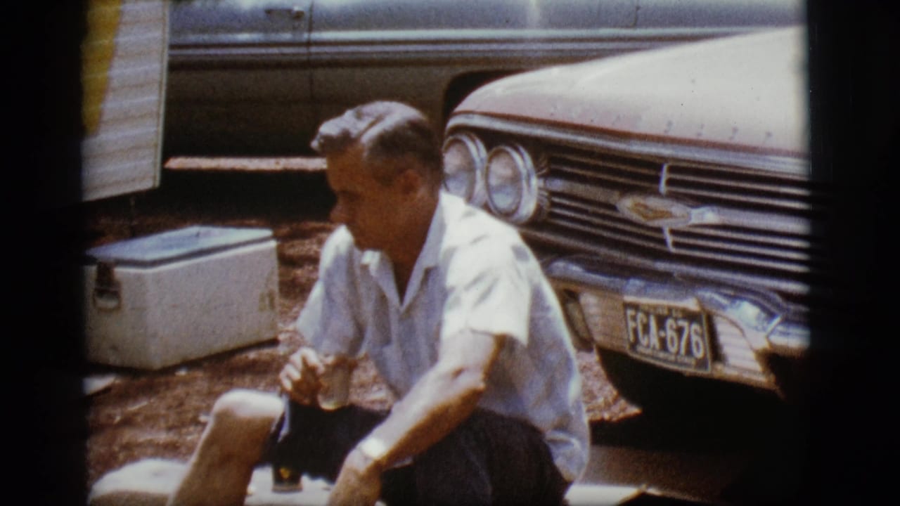 Man seated beside a 1960s Chevrolet with a white cooler nearby.
