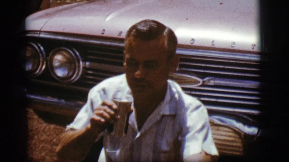The seated man remains close to the Chevrolet front end, holding a can as the handheld 8mm frame shifts slightly.