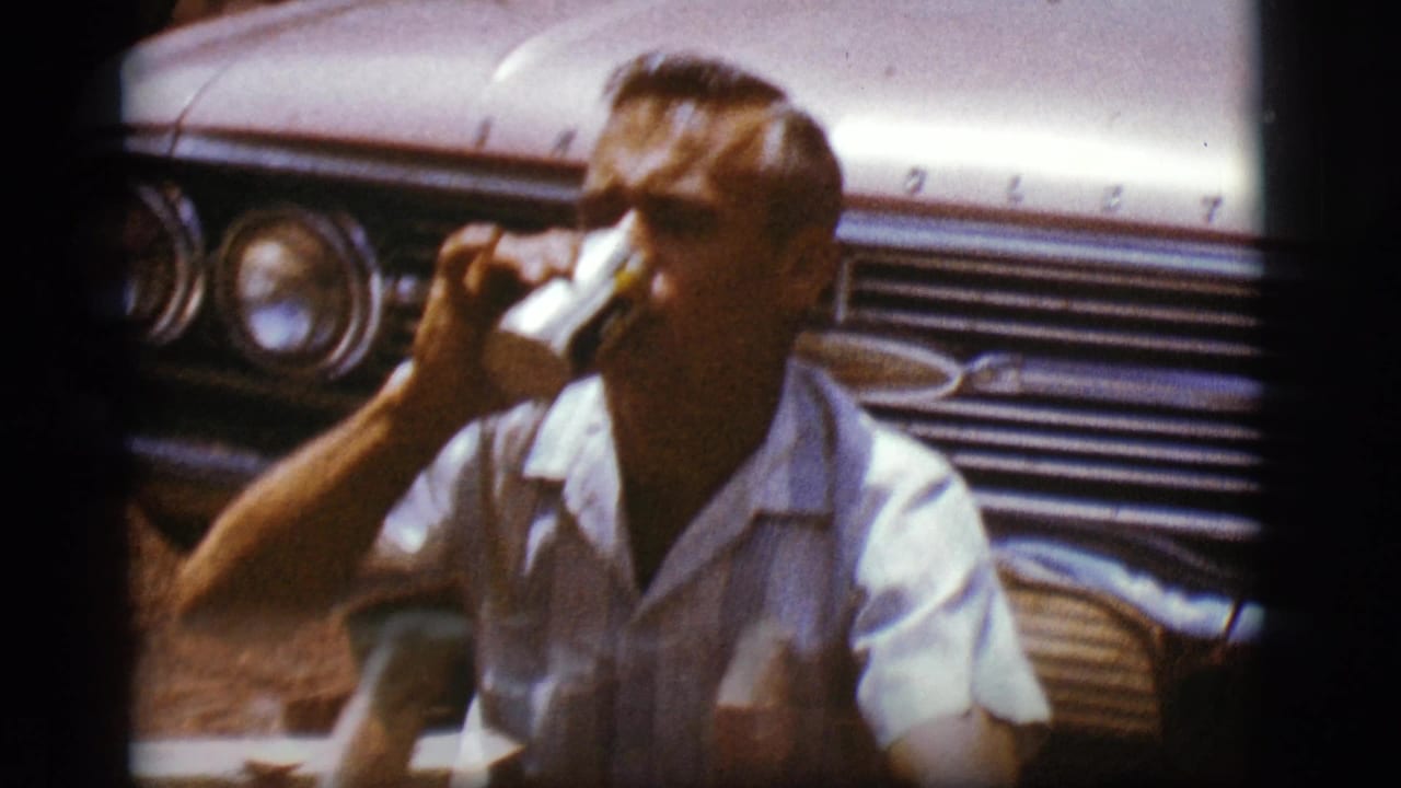 Man in a light shirt drinks from a can beside the front of a vintage Chevrolet.