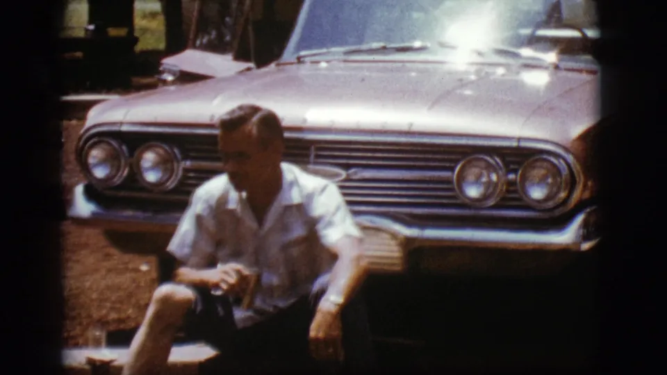 The 8mm frame shows a seated man before a 1960s Chevrolet, with the chrome grille, headlights, and bright hood filling the background.