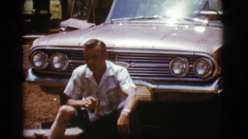 A man sits with a can in front of a Chevrolet during a 1968 Flagstaff home-movie moment, with strong sunlight on the car hood.