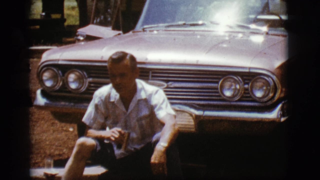 Man seated in front of a light-colored classic sedan, holding a drink in bright sun.