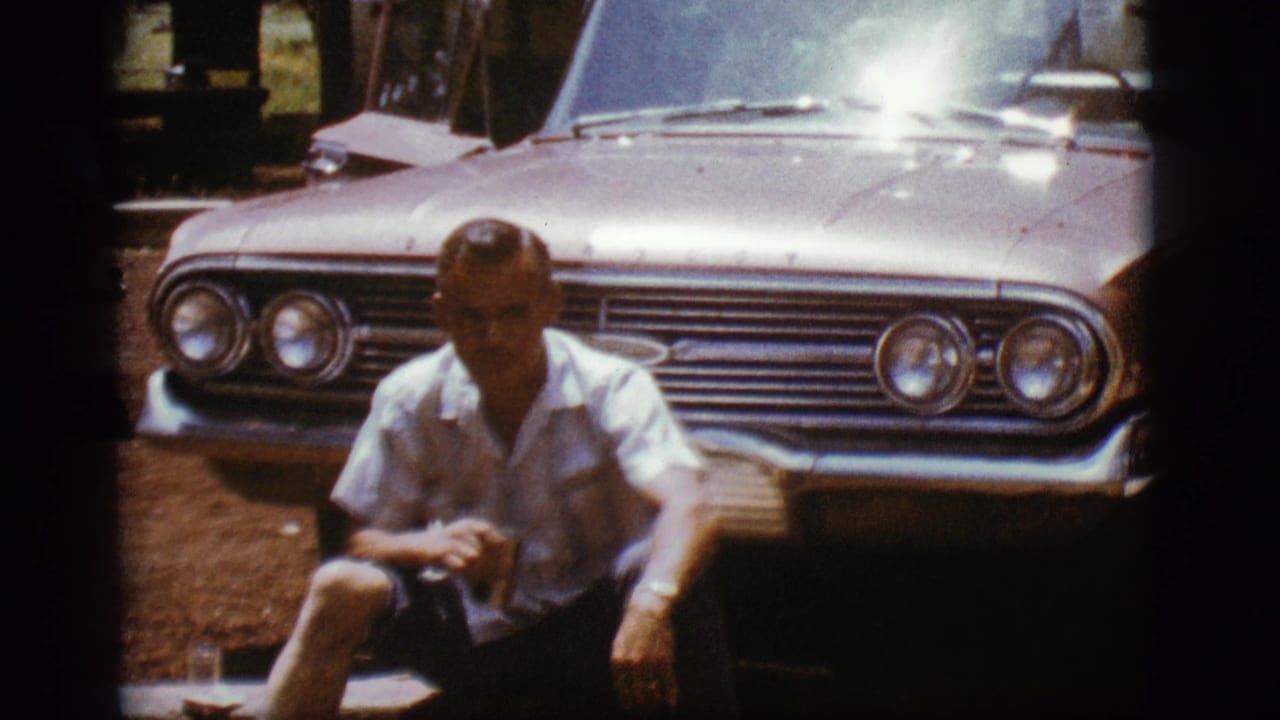 The man remains seated before the 1960s sedan in bright outdoor light, with glare on the hood and windshield.