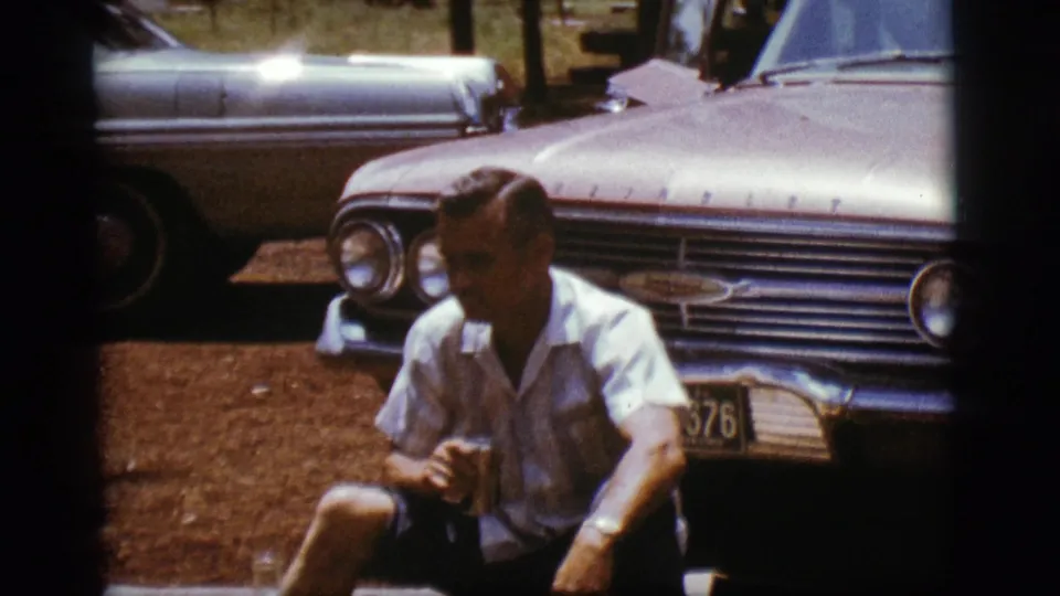 A slightly shifted 8mm frame shows the seated man beside the front end of the sedan, with another older car partly visible behind him.