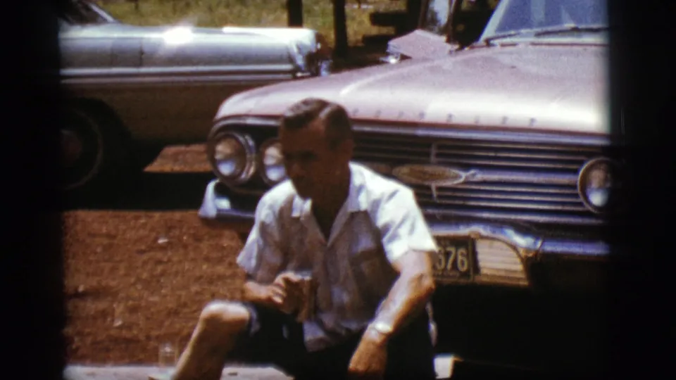 The final frame shows the man seated in front of the sedan's chrome grille and license plate, with another car partly visible in the background.