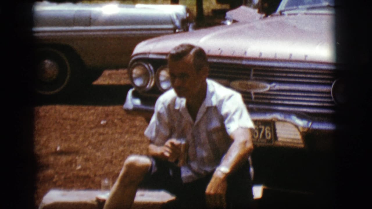 Adult man seated with a can in front of chrome-fronted parked cars.