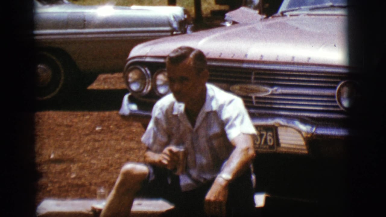 A seated adult man holds a can beside a chrome-grilled 1960s car in supplied Flagstaff, Arizona footage from 1968.