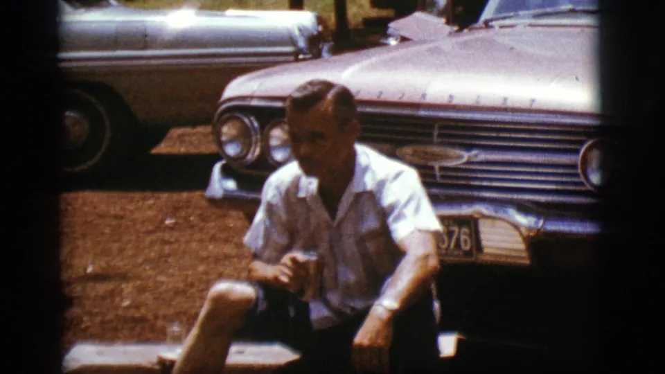 A seated adult man holds a can beside a chrome-grilled 1960s car in supplied Flagstaff, Arizona footage from 1968.
